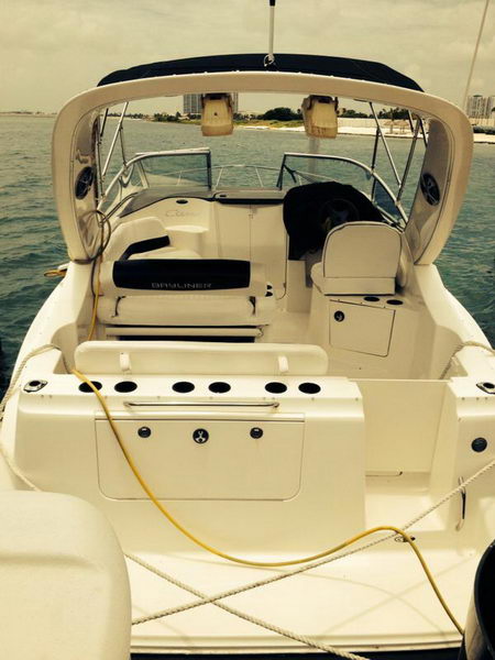 Aft cockpit of a Bayliner cabin cruiser with a helm, bimini and dark canvas cover, a swim platform with cup holders and a yellow hose, viewed against a grey overcast sea with a distant city skyline.