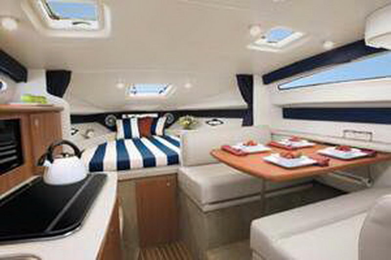 Bright interior cabin of a Bayliner cruiser, likely a stock/manufacturer photo, showing a V-berth with blue and white striped bedding, a galley with a kettle on a stovetop and a dinette table set with red placemats and wood trim.