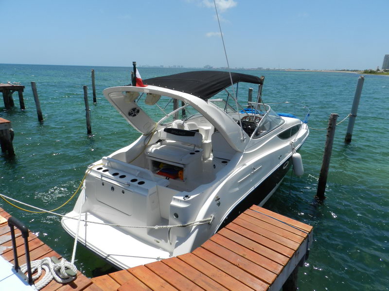 Aft view of a white Bayliner cabin cruiser with a black bimini and Mexican flag moored at a wooden dock on turquoise-green water with wooden pilings and the Cancun skyline on the horizon under a bright sky.