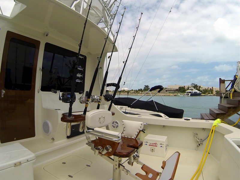 Cockpit of a sportfishing boat looking forward, multiple trolling rods in holders, a fighting chair, the salon door and bait-prep station. Marina water and a megayacht visible to the side; partly cloudy.