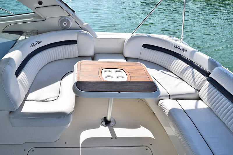 Aft cockpit of the Sea Ray with white U-shaped lounge seating, dark piping and a teak table with built-in cupholders on a pedestal, the green water visible just beyond the transom.