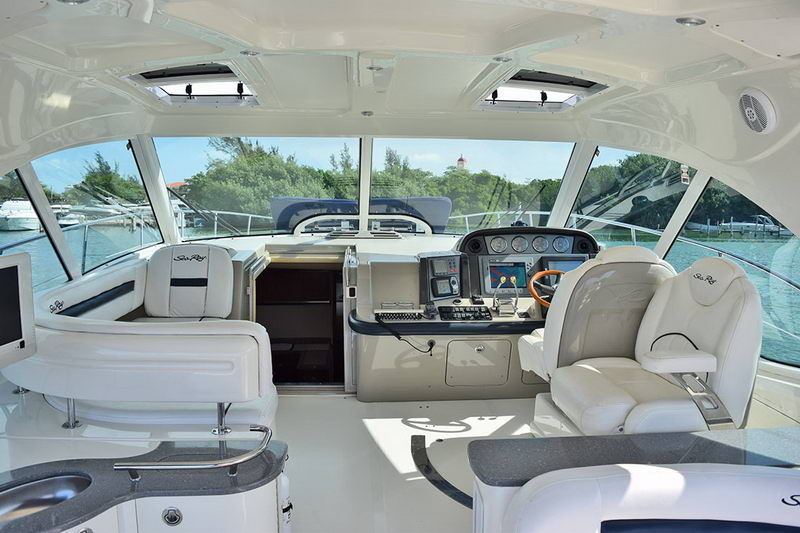 Interior helm and cockpit of the Sea Ray under a hardtop, with white leather captain and companion seats, a dashboard of gauges, twin chartplotters, a wood-rimmed wheel and wraparound windows.