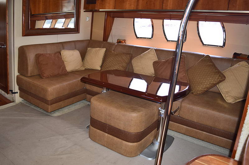 Salon interior with a tan and brown leather L-shaped sofa, accent pillows, a glossy wood cocktail table and a row of portholes along the hull side. Warm cabin lighting.