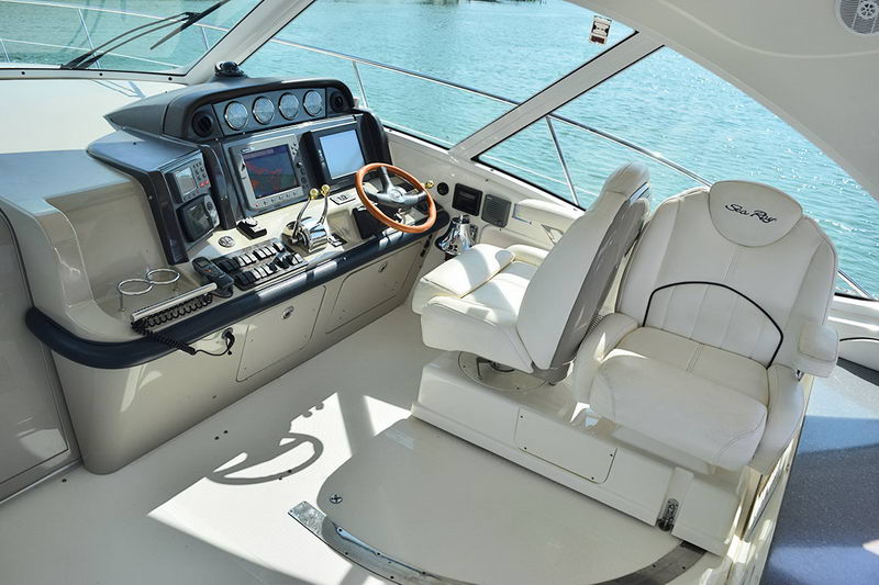 Lower helm station with white leather seats, a wood-rimmed wheel, banks of round gauges and chartplotter screens set into a dark dashboard, viewed beside the windshield.