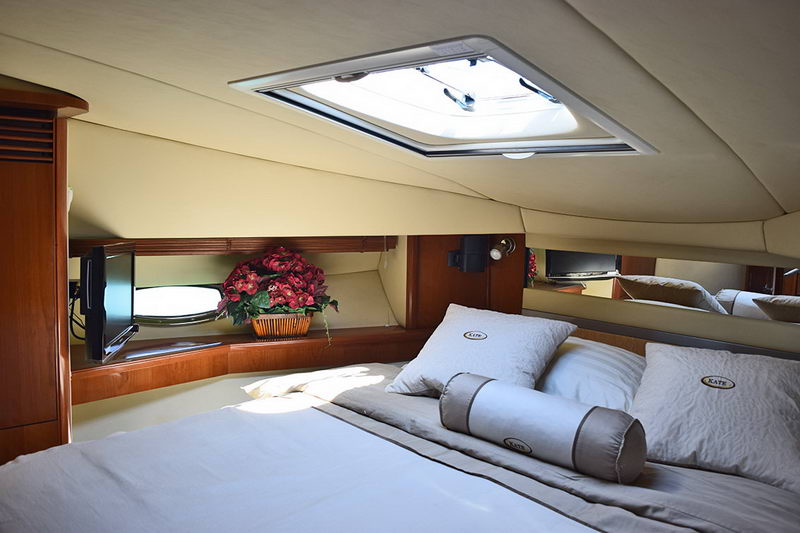 Master cabin with a bed dressed in grey bedding and bolster pillows monogrammed 'Kate', an overhead hatch with daylight, a small TV, wood cabinetry and a basket of red flowers.