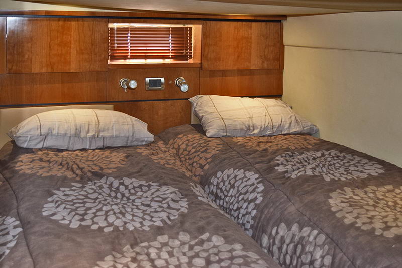 Guest cabin bed made up with brown and grey floral-patterned bedding against a wood headboard fitted with reading lights and a blind-covered window. Cozy enclosed berth.