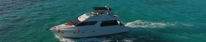 Narrow banner aerial of a white flybridge motor yacht cruising on bright turquoise water with several people aboard on the upper deck.