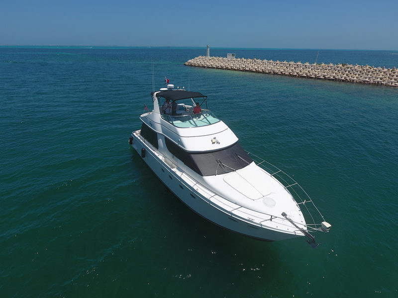 Aerial view of a white flybridge motor yacht with a lone person at the helm, anchored on turquoise water near a rocky breakwater under a clear sky.