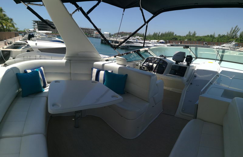 Flybridge of a yacht with white leather L-seating, blue accent pillows, a folding table and the helm console on the right, looking over a marina with other yachts.
