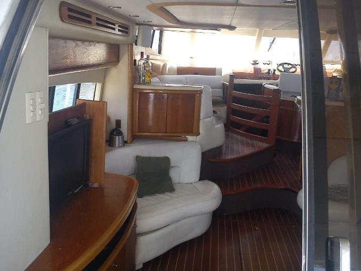 Salon interior viewed from the entry, white leather seating, glossy wood cabinetry and bar, a flat-screen TV, an interior helm with wheel, and a teak-and-holly sole.