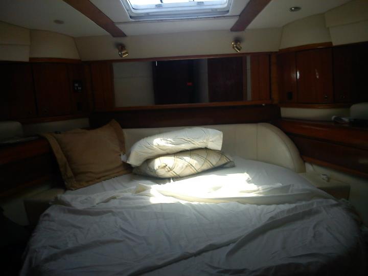 Dimly lit cabin with a made bed in white bedding and pillows, glossy wood trim and cabinets, an overhead hatch letting in a shaft of sunlight.