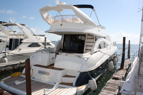White flybridge motor yacht moored at a dock, shown from the side with strong backlight glare off the sea. Flybridge with radar arch and a wood staircase to the cockpit visible. Sunny conditions.