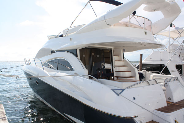 View from a yacht's flybridge looking aft over a white cushioned bench seat and a small table toward the open sea. Stainless rails frame the scene; scattered clouds and bright horizon beyond.