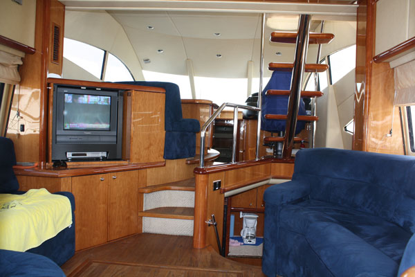 Interior salon and dinette with blue suede armchairs, a CRT television and VCR in a wood console, glossy cherry cabinetry and a curved staircase. Cream headliner and wood flooring; lived-in look.