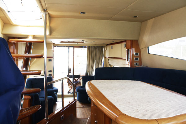 Interior salon dinette with a blue cushioned settee, a varnished wood table with woven inlay top, beige panelling and a wood staircase. Sliding doors at the rear lead to the cockpit; daylight throughout.