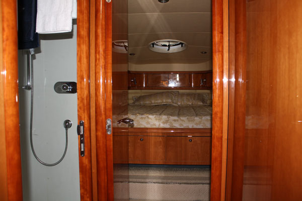 View through a glossy cherry-wood doorway into a cabin showing a made double bed with patterned cover, overhead light and storage drawers. An ensuite with a handheld shower is visible on the left.