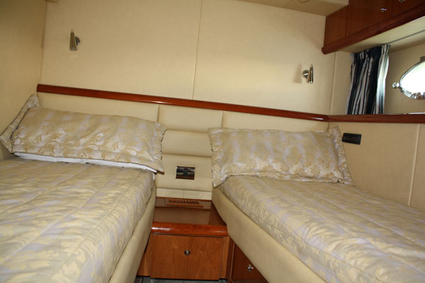 Guest cabin with two single beds arranged in a V, dressed in cream patterned bedding with matching pillows, cherry-wood trim, reading lights and a small mirror. Tidy and well lit.