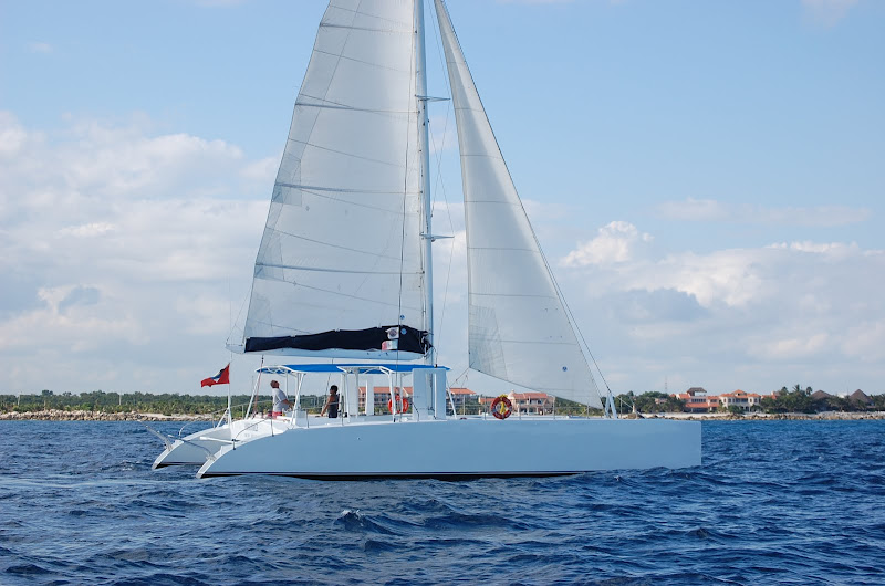 A white sailing catamaran under full white sails with a few people aboard on the foredeck, cruising on deep blue water with a low green coastline and resort buildings in the distance under a bright sky.