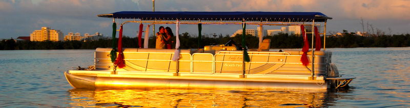 Banner-format photo of a pontoon party boat on a calm lagoon at sunset, with a blue canopy decorated with Christmas garlands and a couple embracing aboard. Warm golden light reflects on the water.