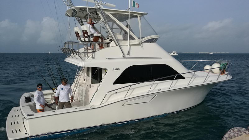 White Cabo sportfishing yacht with tuna tower and outrigger rods, several crew aboard the cockpit and tower, underway on blue water with cloudy skies and the Mexican flag flying from the tower.