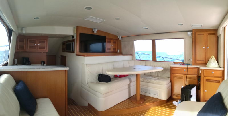 Interior salon of a sportfish yacht (panoramic) with cream leather dinette and sofas, light wood cabinetry, a flat-screen TV, galley with coffee maker, teak sole, and windows showing open sea.