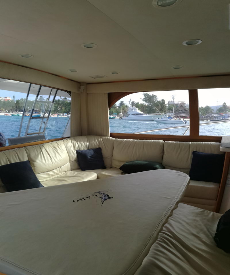 Interior of a sportfish yacht salon with a large cream leather wraparound settee and dinette table embroidered 'CABO', cushions, and big windows showing a marina and another yacht passing outside.