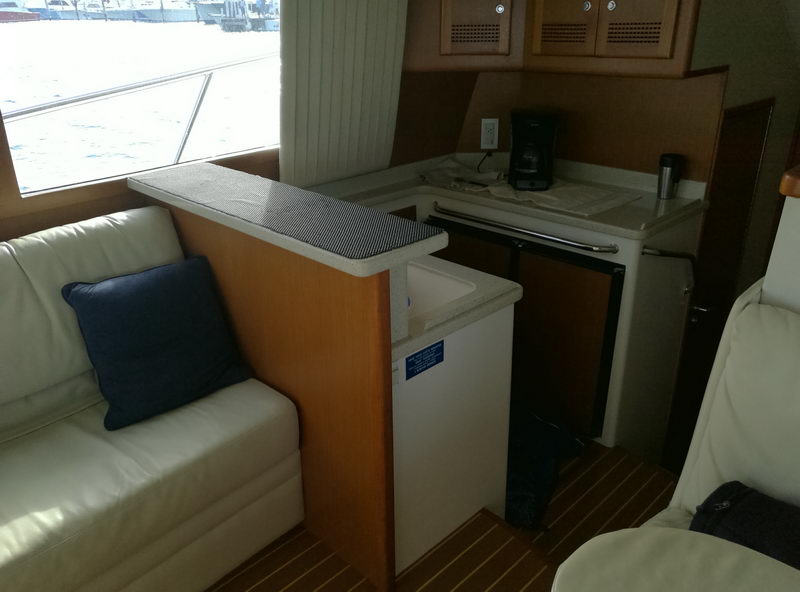 Interior of a sportfish yacht showing a small galley/bar area with light wood cabinetry, white counters, a coffee maker, mini-fridge, cream leather seat with a navy pillow, and teak flooring.
