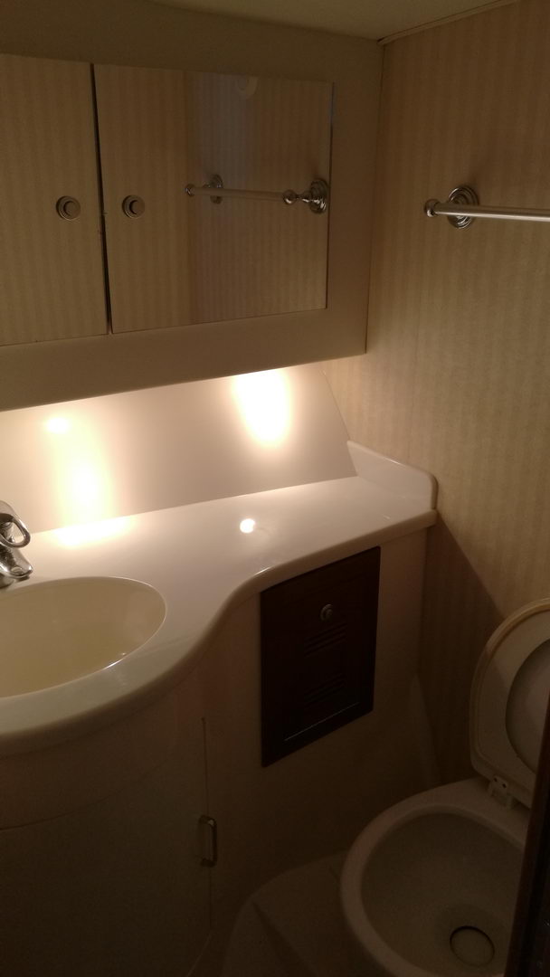 Yacht bathroom (head) with a white molded vanity and oval sink, mirrored cabinet, a dark wood access panel, cream striped wallpaper, warm lighting and a marine toilet at right.