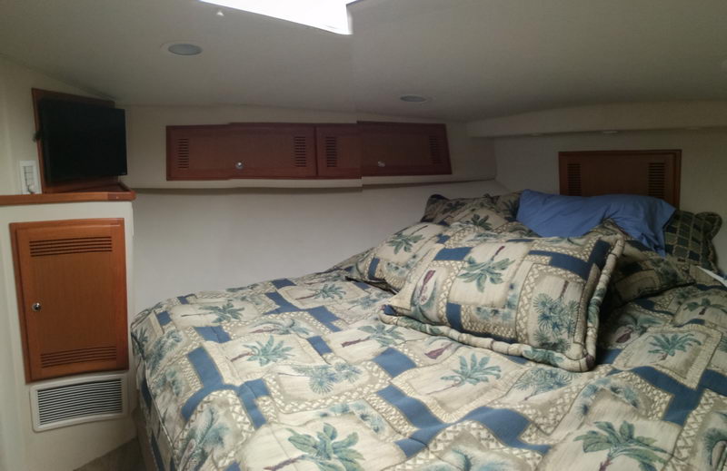 Forward cabin of a sportfish yacht with a V-berth made up in palm-tree patterned bedding and a blue pillow, light wood lockers and a flat-screen TV mounted on the bulkhead, overhead hatch above.
