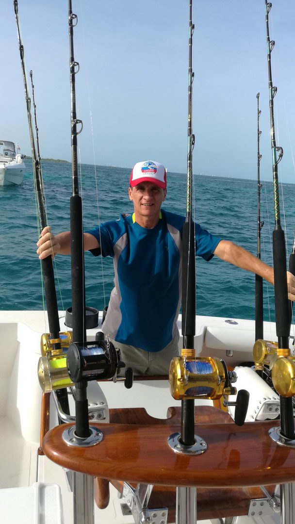 A smiling man in a blue shirt and red cap stands behind a row of heavy trolling rods in rod holders on the cockpit of a sportfishing boat, with gold and chrome reels and turquoise sea behind him.