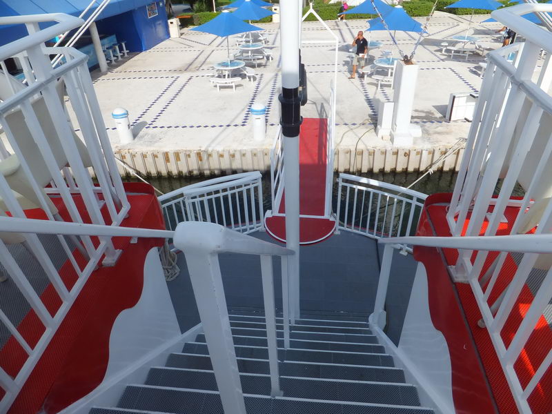 Vista por la escalera roja del Cancun Queen hacia la terraza del muelle con sombrillas azules en la marina de Cancún