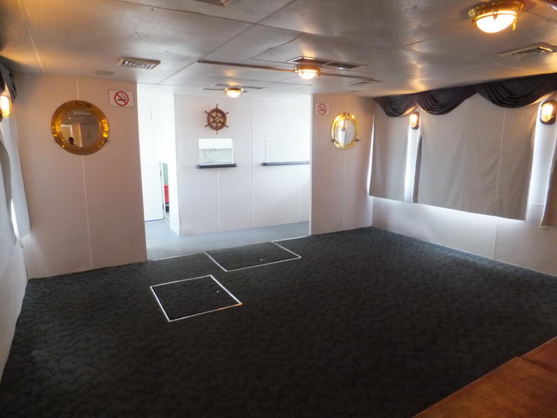 Salón interior del Cancun Queen con decoración de timón, ojos de buey dorados y barra, barco privado de fiesta en Cancún