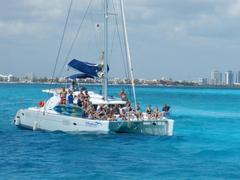 White Lagoon 'Albatros' sailing catamaran loaded with many guests on deck, anchored in vivid blue-and-turquoise water with a kayak aboard, Cancun hotel-zone skyline in the background under a partly cloudy sky.