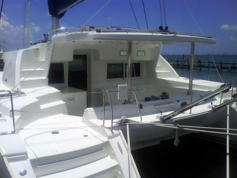 Stern-quarter deck view of a white Lagoon catamaran moored at a dock, showing the cockpit, coachroof windows, stainless arch and rails and aft trampoline area, with the harbor and distant shoreline beyond under a blue sky.