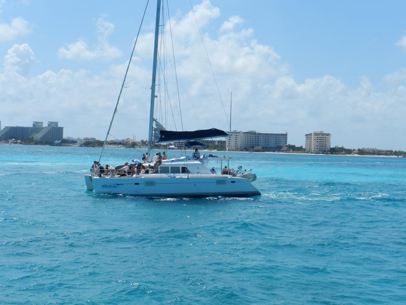 White Lagoon 'Albatros' catamaran motoring in blue Caribbean water with guests aboard under a furled blue bimini, the Cancun hotel zone of high-rise resorts lining the shore behind under a sunny sky.