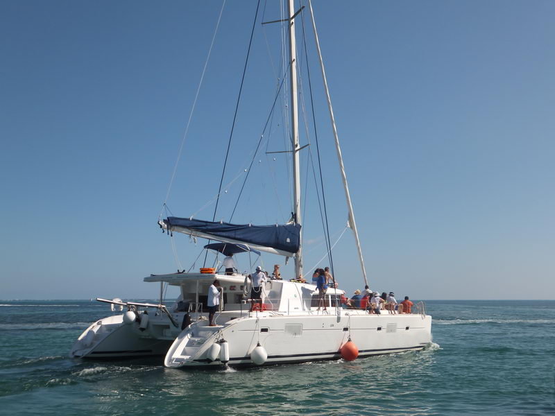 Three-quarter stern view of a large white Lagoon catamaran underway on calm turquoise-blue water with several guests on the aft deck, mast and furled mainsail prominent, distant low shoreline under a clear sky.
