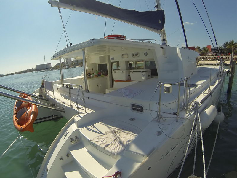 Bow-to view of a large white Lagoon catamaran moored at a dock, showing the foredeck with trampolines, coachroof windows, cockpit, an orange life ring and laid-out towels, with resort buildings behind on a sunny day.