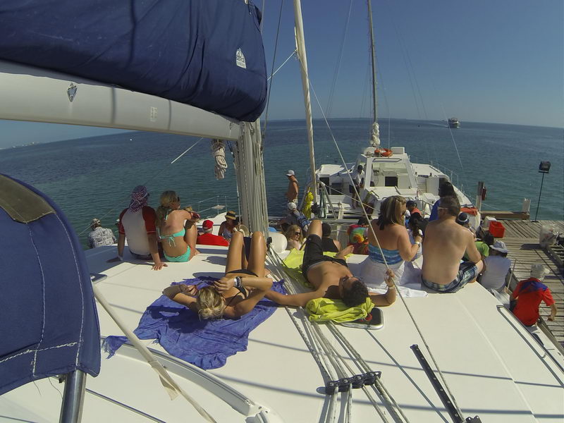 On-deck view from the coachroof of a large catamaran with many guests in swimwear sunbathing and sitting on the white sunpads and side decks, the boat docked beside a pier with open sea and another boat in the distance.