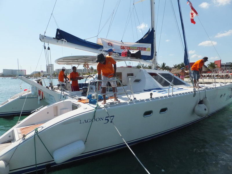 Bow-to view of a large white 'Lagoon 57' catamaran at a dock with several crew in orange shirts working on deck and a promotional cancunboozecruise banner on the bimini, moored in turquoise water with beach and palms behind.