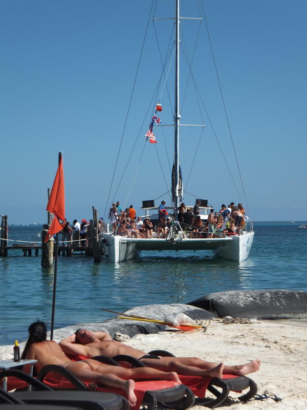 View from a sandy beach toward a white catamaran packed with guests moored at a dock in calm turquoise water, an orange flag and sunbathers on red loungers in the foreground under a clear blue sky.
