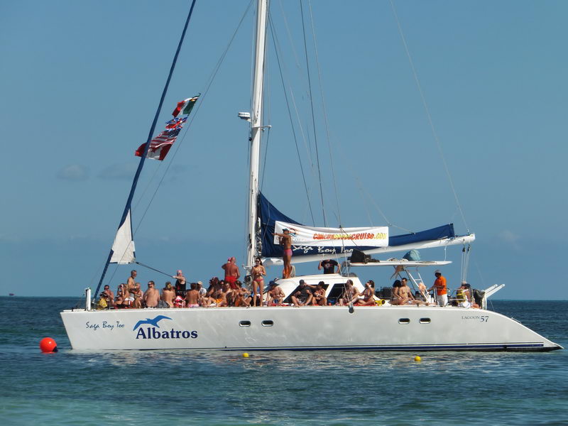 White Lagoon 57 sailing catamaran branded 'Albatros / Saga Bay Too' anchored offshore in turquoise water, crowded with many passengers in swimwear sunbathing on deck. Mexican and other flags fly from the rigging on a sunny clear day.