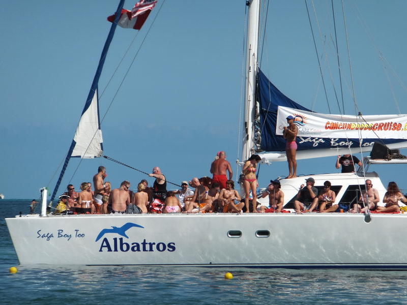 Close-up of the white 'Albatros / Saga Bay Too' catamaran bow packed with sunbathing passengers in swimwear. A banner advertising a Cancun booze cruise hangs from the rigging on a sunny day with calm sea.