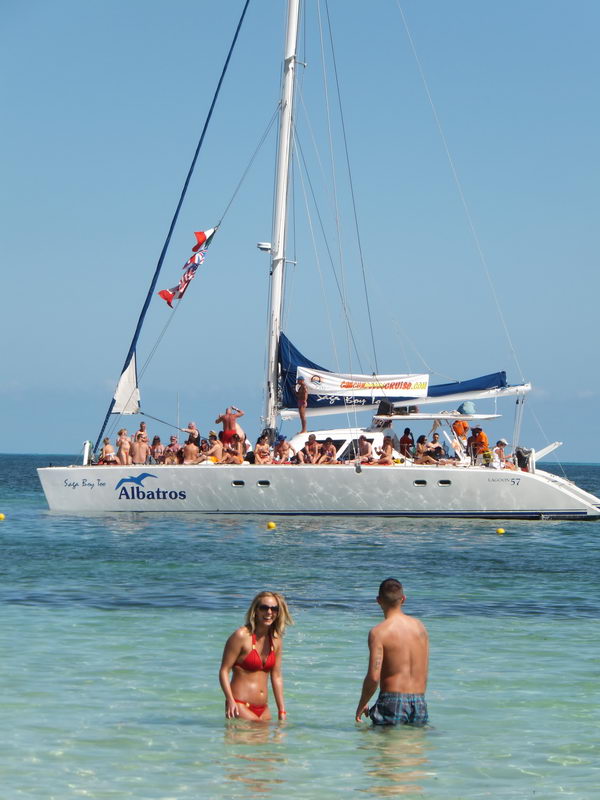 Vertical shot of the 'Albatros' Lagoon 57 catamaran full of passengers anchored offshore, with a couple (woman in red bikini and a man) standing in shallow turquoise water in the foreground on a sunny day.