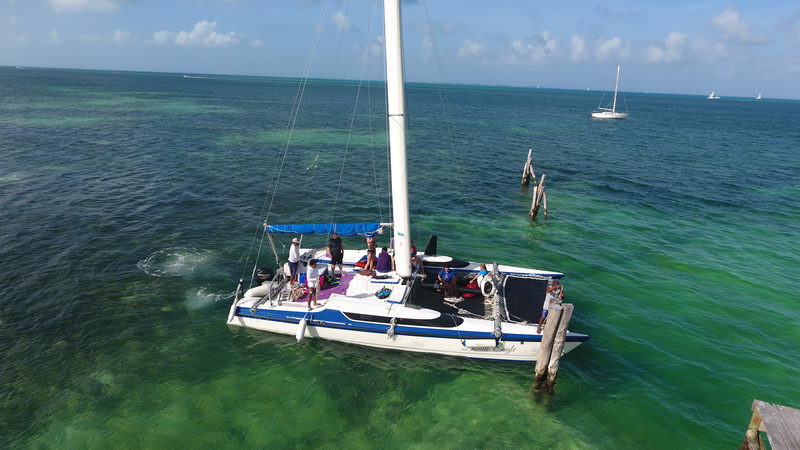 Aerial of the 'Double Eagle' catamaran with passengers backing into a marina dock, many sportfishing boats moored alongside, turquoise water and Cancun shore behind, sunny.