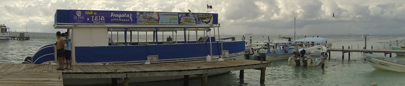 Wide panoramic dock photo of a blue double-decker tour pontoon boat labeled 'Fragata' moored at a wooden pier, overcast sky. People stand on the dock; small boats and other vessels float alongside in a harbor on Isla Mujeres.