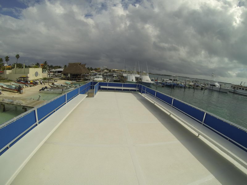 Empty white upper sun deck of the Fragata tour boat looking forward, with blue safety railings and a textured non-slip floor. Overcast cloudy sky overhead; a beach with palapas, parked boats and waterfront buildings to the left.