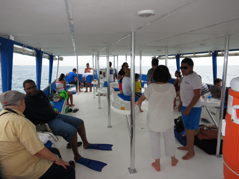 Daytime interior upper deck crowded with passengers, adults and children seated on blue cushions and standing. Snorkel fins on the floor, an orange water cooler, open-sea horizon visible on all sides, sunny.