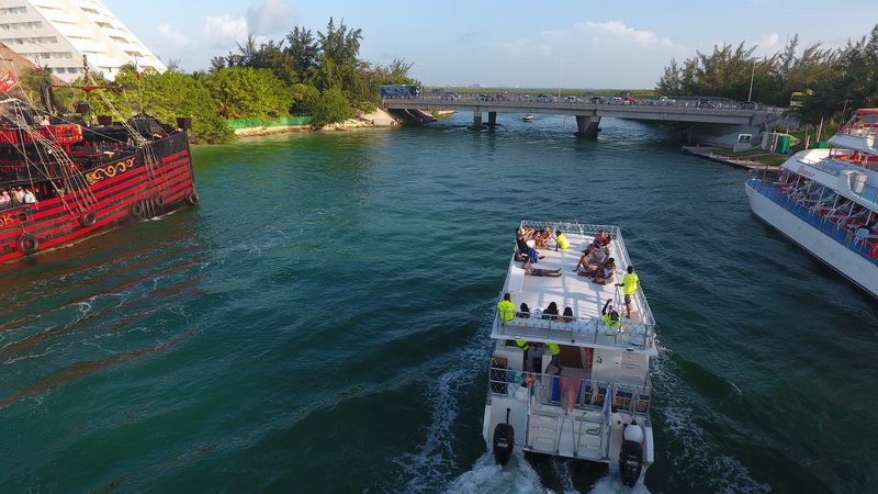 Aerial rear view of the Fragata boat cruising through a channel beneath a road bridge. A red pirate-ship vessel and another tour catamaran are nearby; green water, people on deck, twin outboard wakes, sunny.