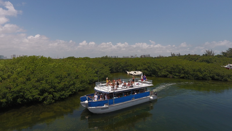 Aerial view of the Fragata cruising through a mangrove lagoon, people on the upper deck. Lush green mangroves line the channel with the distant Cancun skyline under a blue sky.