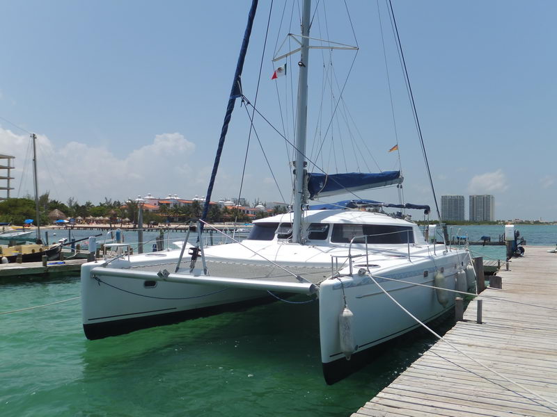 White sailing catamaran moored bow-to at a wooden dock on a sunny day, mast and furled mainsail upright. Turquoise-green water, with a beach and resort buildings behind.